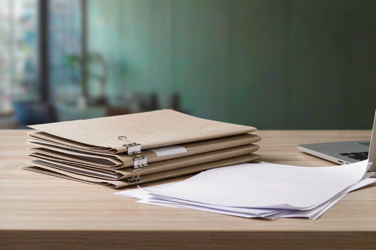 A stack of file folders and documents on a bright desk next to a laptop in soft morning light