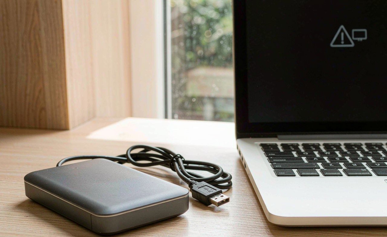 An external portable hard drive and USB cable on a wooden desk next to an open laptop