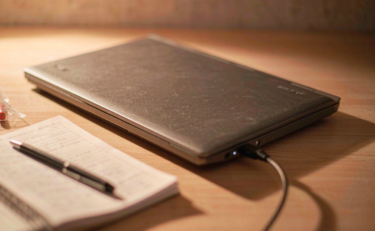 A closed gray laptop on a desk with its charger plugged in, a small white power LED glowing beside a notebook and pen