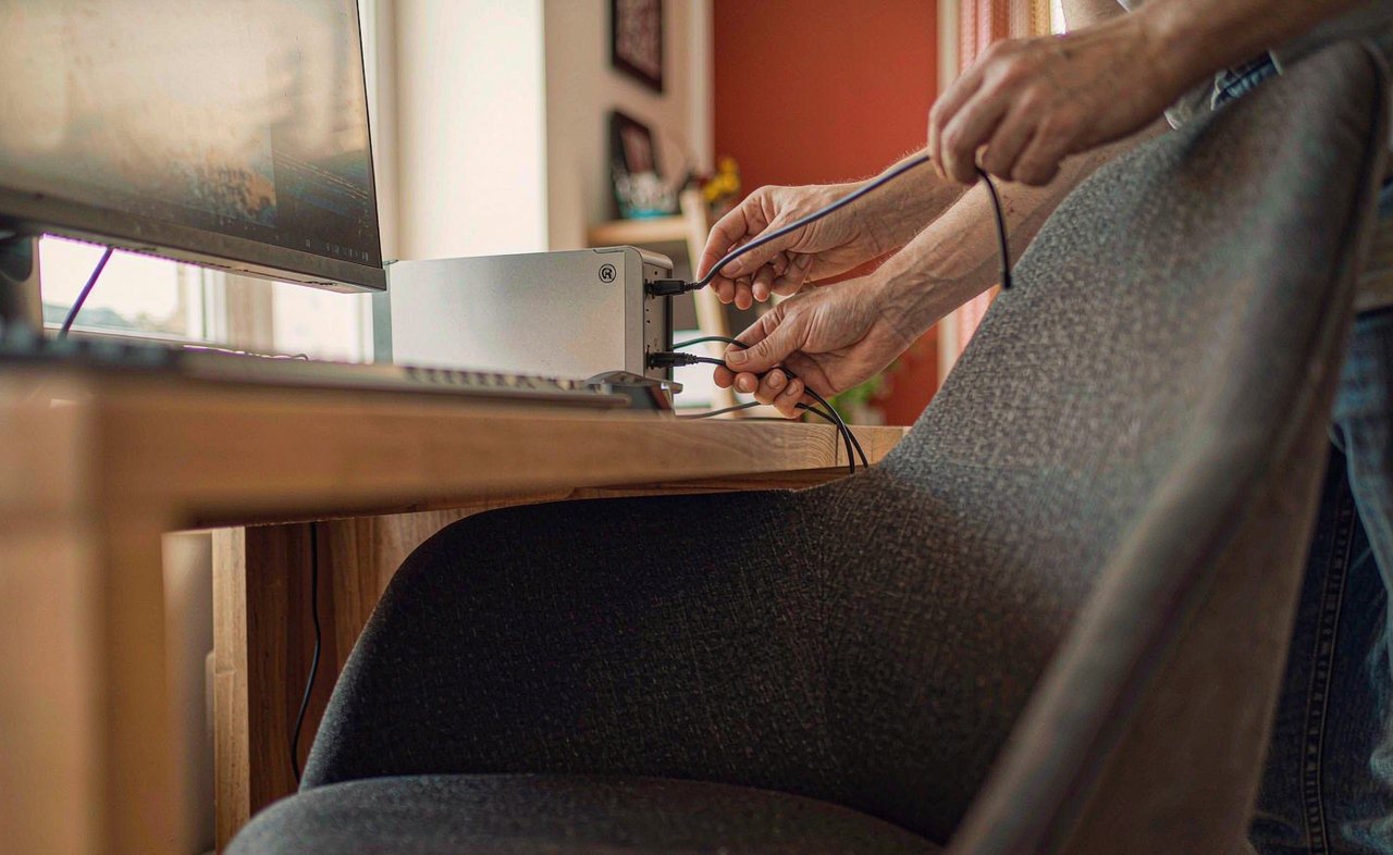 A person plugging a headphone jack into the audio port on the back of a desktop computer tower under a wooden desk