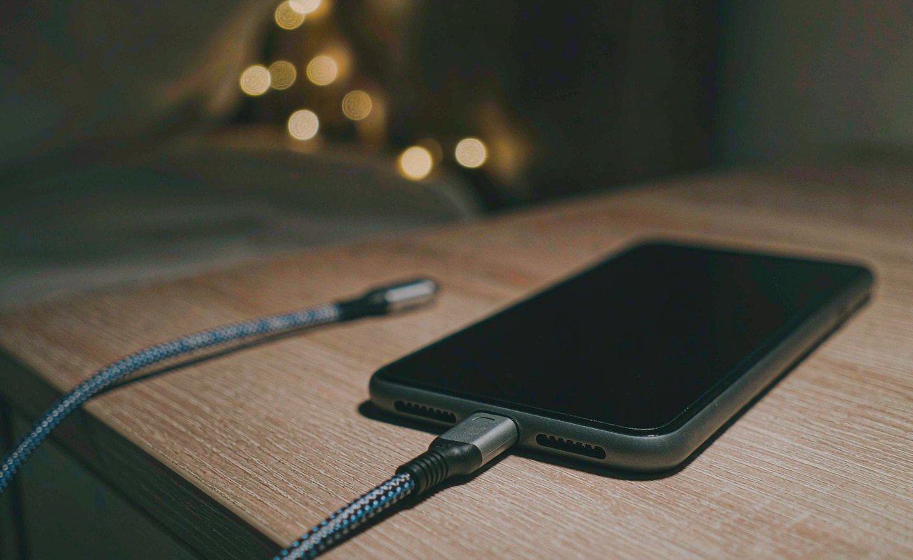 A smartphone on a wooden nightstand connected to a charging cable in soft bedside lamp light