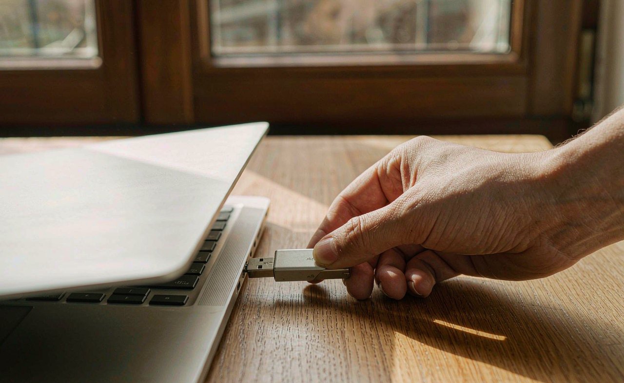 A hand plugging a compact metal USB flash drive into the side port of a silver laptop on a wooden desk in warm morning light
