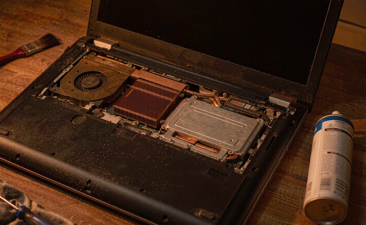 A laptop on a wooden desk with its cooling fan visible through a partially opened bottom panel, a small brush and compressed air can beside it, warm workshop lighting