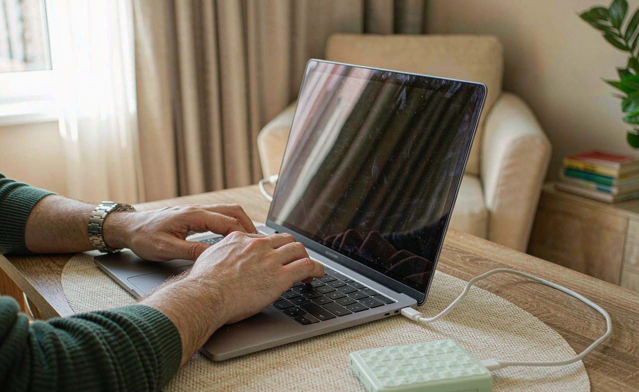 Hands typing on a laptop keyboard in daylight next to a connected external hard drive on a bright desk