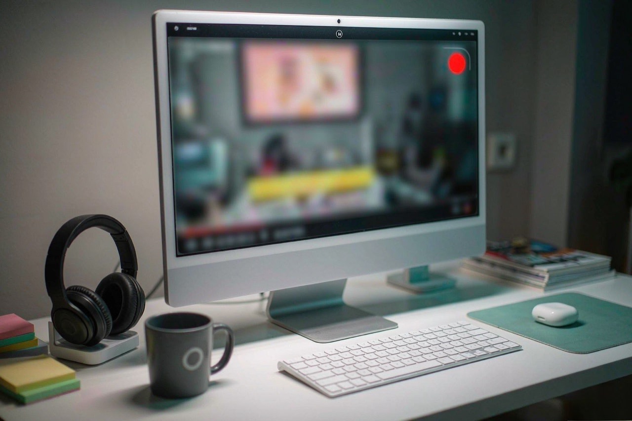 A desktop monitor showing a recording interface with a red record button, headphones and keyboard on the desk