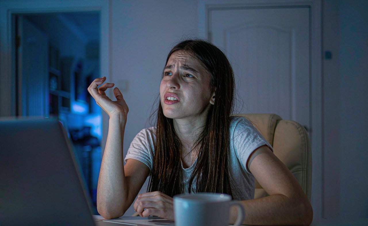 A frustrated woman leaning back at a home desk looking at a laptop screen filled with overlapping pop-up windows, cool blue evening lighting from the monitor