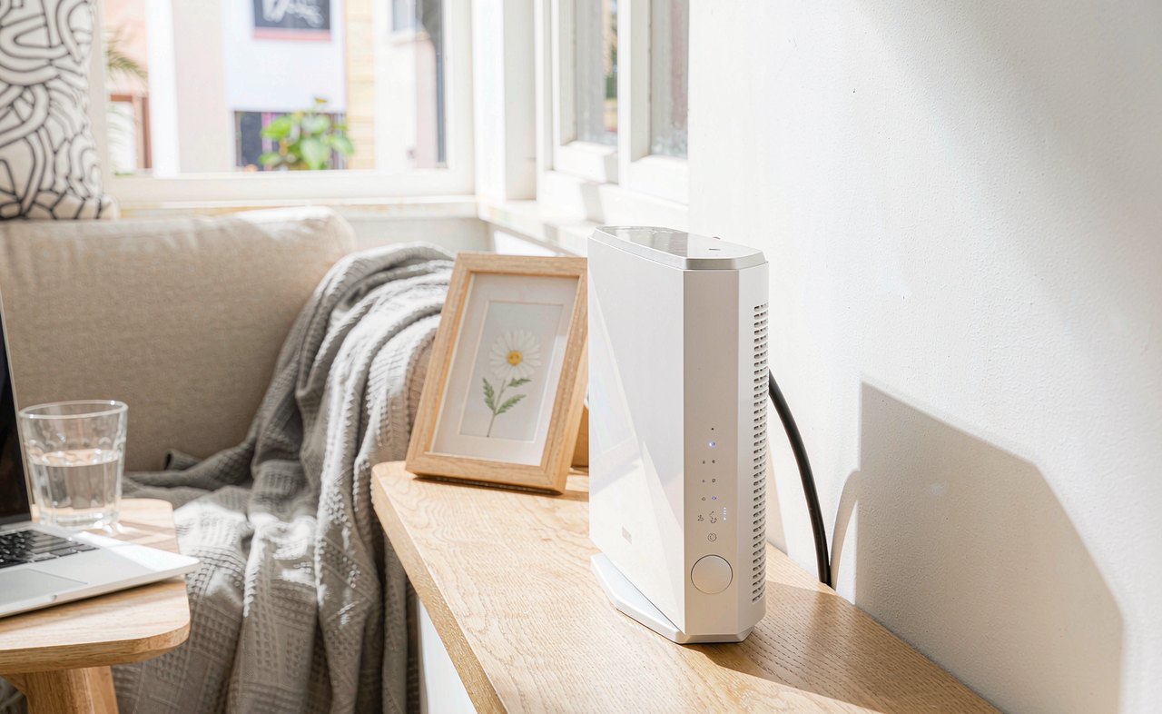 A white Wi-Fi router with glowing indicator lights on a wooden shelf beside an open laptop in a modern living room