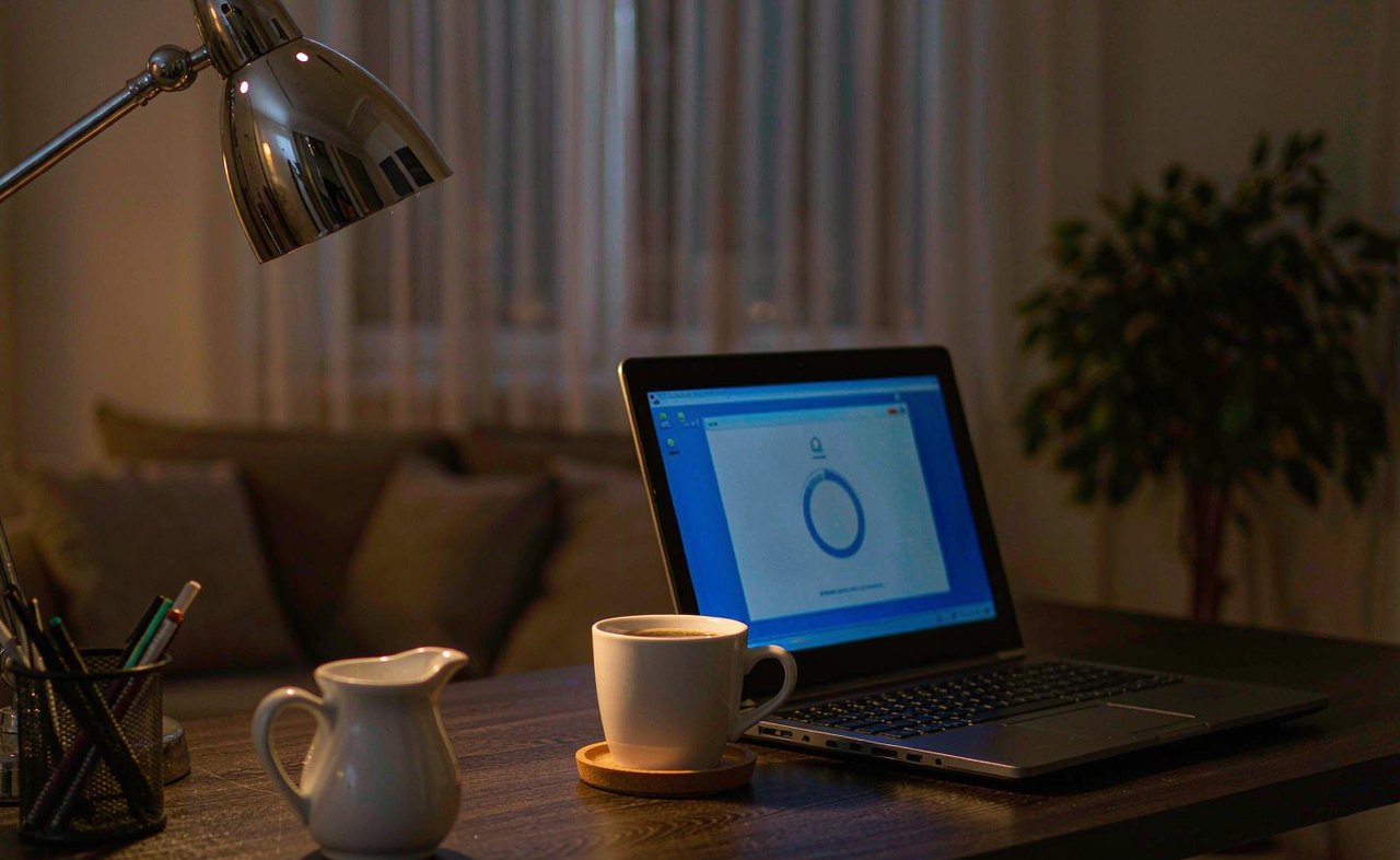 A laptop on a desk at night showing a blue update progress screen, lit softly by a warm desk lamp